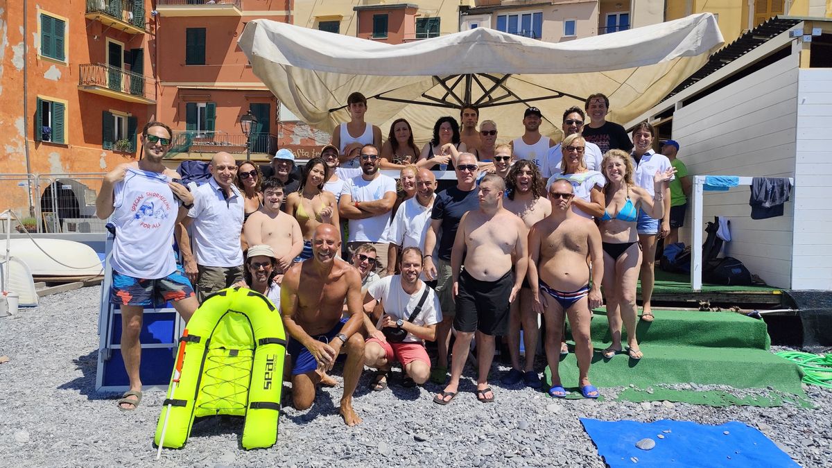 Allievi e istruttori del Centro in posa in spiaggia a Camogli, indossando magliette bianche con il logo del Centro Subacqueo. Sullo sfondo si vedono edifici colorati in stile mediterraneo e il mare.